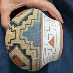 Op de afbeelding: Een ronde keramische kom met een textuurrijk oppervlak en geometrische patronen in beige, zwart en grijs. De rand is glad en wit, versierd met ingesneden geometrische lijnen en een driehoeksvorm die de terracotta klei eronder blootlegt.