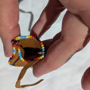 Puede incluir: Peque&ntilde;o bolso de cuero marr&oacute;n con un borde de cuentas. Las cuentas tienen un patr&oacute;n de azul, blanco, amarillo, rojo y negro. Una fina correa de cuero se extiende desde la parte inferior del bolso. El bolso se sostiene entre dos dedos.