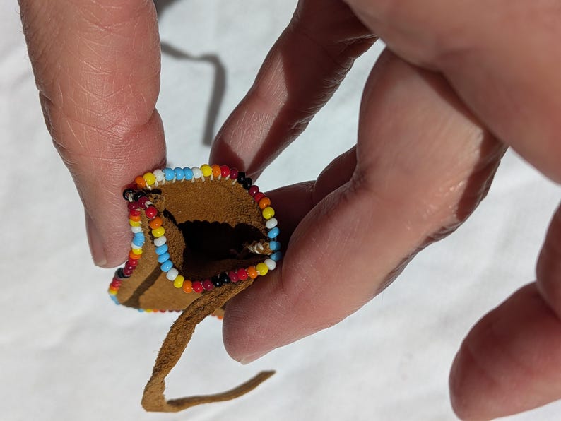 Puede incluir: Peque&ntilde;o bolso de cuero marr&oacute;n con un borde de cuentas. Las cuentas tienen un patr&oacute;n de azul, blanco, amarillo, rojo y negro. Una fina correa de cuero se extiende desde la parte inferior del bolso. El bolso se sostiene entre dos dedos.