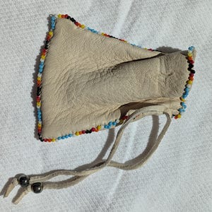 Puede incluir: Una bolsa de cuero color crema con un borde de cuentas rojas, amarillas, azules y negras. La bolsa tiene un cierre de cord&oacute;n con dos cuentas de color gris oscuro. La bolsa est&aacute; sobre una superficie blanca texturizada.