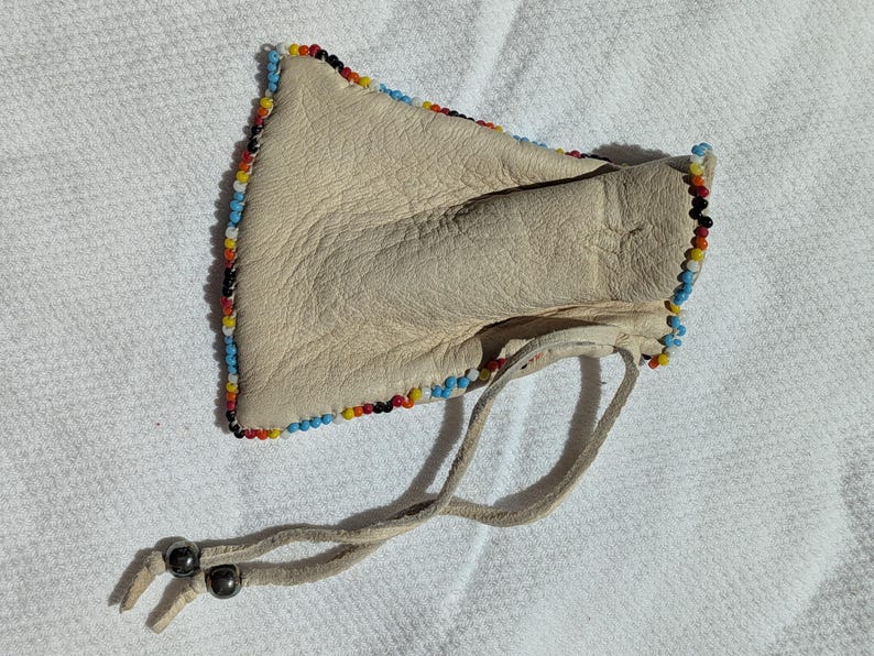 Puede incluir: Una bolsa de cuero color crema con un borde de cuentas rojas, amarillas, azules y negras. La bolsa tiene un cierre de cord&oacute;n con dos cuentas de color gris oscuro. La bolsa est&aacute; sobre una superficie blanca texturizada.