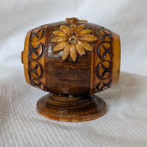 May include: A wooden barrel-shaped decorative item with a flower on top. The item is brown with lighter wood accents and carved designs. It sits on a small, round base.