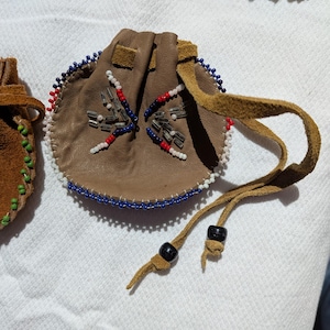 Puede incluir: Una peque&ntilde;a bolsa redonda de cuero marr&oacute;n con cierre de cord&oacute;n. La bolsa est&aacute; decorada con cuentas azules, rojas y blancas alrededor del borde y en un patr&oacute;n de estrella. El cord&oacute;n tiene cuentas negras.