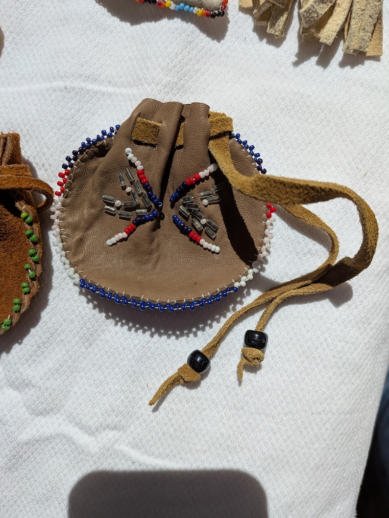Puede incluir: Una peque&ntilde;a bolsa redonda de cuero marr&oacute;n con cierre de cord&oacute;n. La bolsa est&aacute; decorada con cuentas azules, rojas y blancas alrededor del borde y en un patr&oacute;n de estrella. El cord&oacute;n tiene cuentas negras.