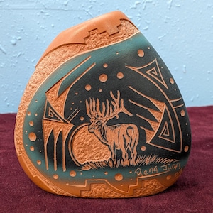 以下が含まれることがあります： 手彫りの、涙の形をした陶器の花瓶。詳細なヘラジカのシーンが描かれています。花瓶は、質感のあるオレンジブラウンの外観と、ヘラジカのイラストが描かれたティールと黒の内装が特徴です。アーティストのサイン、Rena Juan が見えます。