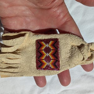 Puede incluir: Una pulsera de cuero beige con un dise&ntilde;o rectangular de cuentas. La secci&oacute;n de cuentas presenta un patr&oacute;n geom&eacute;trico en rojo, amarillo y blanco. La pulsera tiene un borde con flecos y un cierre anudado.