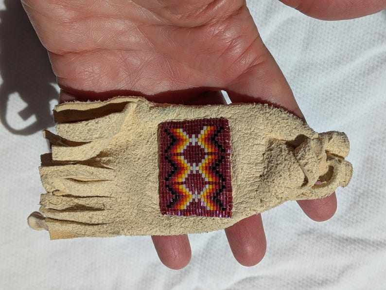Puede incluir: Una pulsera de cuero beige con un dise&ntilde;o rectangular de cuentas. La secci&oacute;n de cuentas presenta un patr&oacute;n geom&eacute;trico en rojo, amarillo y blanco. La pulsera tiene un borde con flecos y un cierre anudado.
