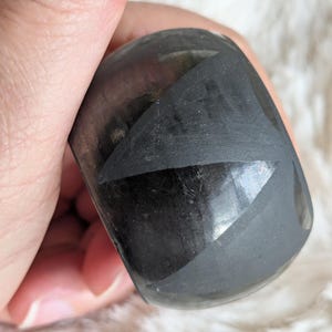 Puede incluir: Una piedra oscura y redondeada con una superficie pulida. La piedra es de color negro intenso con sutiles variaciones tonales. La forma es suave y ergonómica, sostenida en una mano sobre un fondo blanco suave. La piedra parece ser un mineral natural.