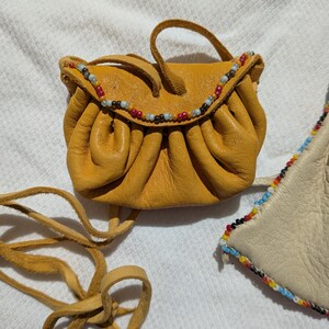 Puede incluir: Bolsa de cuero amarillo mostaza con ribete de cuentas. La peque&ntilde;a bolsa tiene un cierre de cord&oacute;n y un asa corta. Una larga correa de cuero est&aacute; unida a la bolsa. La bolsa est&aacute; sobre una superficie blanca.
