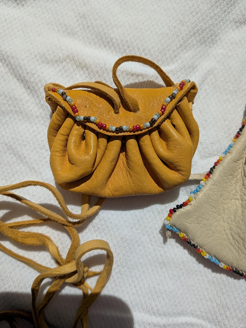 Puede incluir: Bolsa de cuero amarillo mostaza con ribete de cuentas. La peque&ntilde;a bolsa tiene un cierre de cord&oacute;n y un asa corta. Una larga correa de cuero est&aacute; unida a la bolsa. La bolsa est&aacute; sobre una superficie blanca.