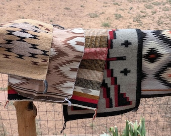 Puede incluir: Una colección de cinco alfombras tejidas rectangulares con patrones geométricos en varias paletas de colores, incluyendo beige, marrón, negro, blanco, rojo y gris. Las alfombras están superpuestas y colgadas sobre una cerca de alambre.