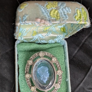 Puede incluir: Un broche de plata y cristal con un camafeo de perfil de mujer en vidrio azul. El broche está en una caja de terciopelo azul claro y verde.