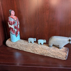 May include: Hand-carved wooden figurines of a Native American woman, two small bears, and a larger bear, all painted in muted colors. The figures are arranged on a natural wood base, creating a rustic and artistic display. The woman wears a red, white, and gray patterned shawl.
