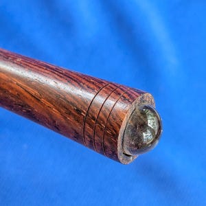 May include: Close-up of a wooden object with a dark brown finish and a rounded end. The wood grain is visible, and there are three carved rings near the end. The end features a dark, spherical element, possibly glass or a similar material.