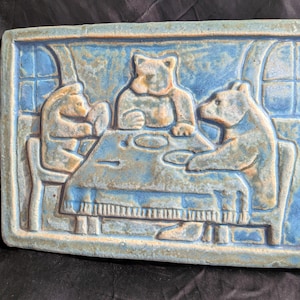 Könnte beinhalten: Rechteckige Keramik-Wandplatte mit einem Relief von drei Bären an einem Tisch. Die Bären sind hellbraun, vor einem marmorierten blauen Hintergrund. Die Szene wird von einem Rahmen mit einem Fenster-ähnlichen Design umgeben.