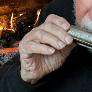 May include: A person plays a silver harmonica with the inscription "MARINE BAND" on it. The person is sitting in front of a fireplace.
