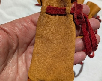Puede incluir: Una pequeña bolsa de cuero hecha a mano en tonos naranja y rojo. La bolsa tiene un cierre de cordón y un fondo con flecos. El cuero tiene un aspecto texturizado. La bolsa se sostiene en una mano.