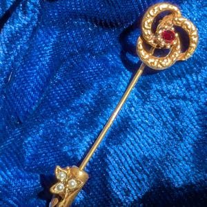 May include: A gold-coloured hat pin with a decorative top featuring a red gemstone and a swirling design. The pin's base has a floral design with three small, white pearls. The pin is set against a blue background.