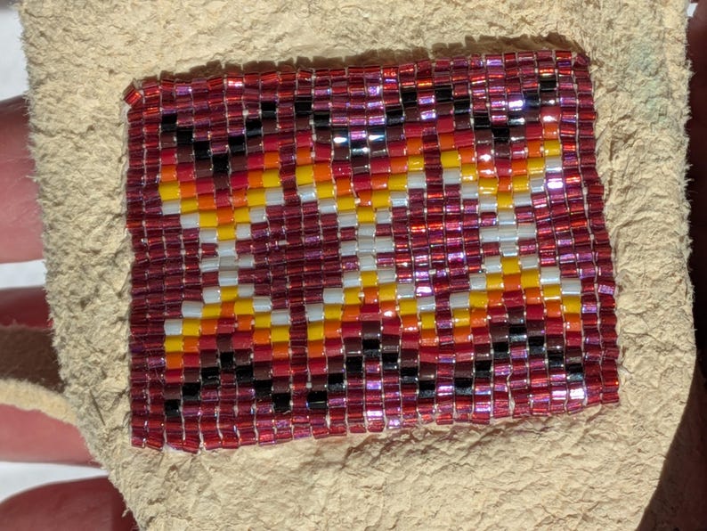 Puede incluir: Un parche rectangular bordado con cuentas con un dise&ntilde;o geom&eacute;trico en tonos de rojo, naranja, amarillo, negro y blanco. Las cuentas est&aacute;n cosidas sobre un respaldo de cuero beige, creando una superficie texturizada. El parche es probablemente un elemento decorativo.