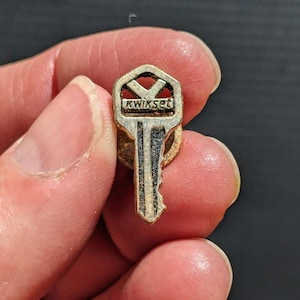 May include: A silver key pin with the word "Kwikset" engraved on the top. The key is held in a hand.