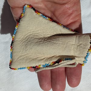 Puede incluir: Bolsa de cuero color crema con borde de cuentas en azul, amarillo, rojo y negro. La bolsa est&aacute; doblada, revelando el interior. El art&iacute;culo se sostiene en una mano.