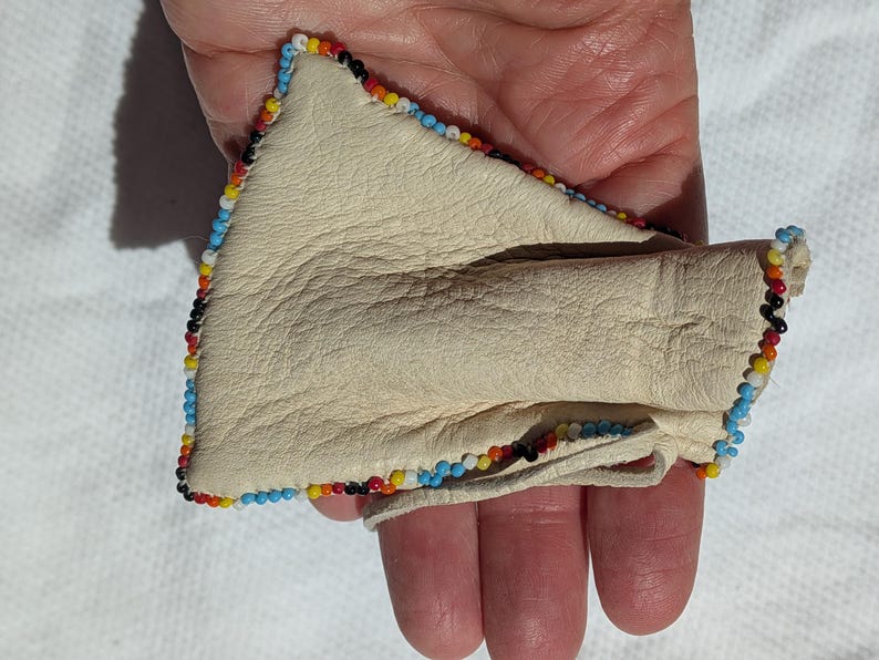 Puede incluir: Bolsa de cuero color crema con borde de cuentas en azul, amarillo, rojo y negro. La bolsa est&aacute; doblada, revelando el interior. El art&iacute;culo se sostiene en una mano.