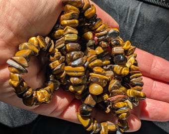 May include: A strand of tumbled tiger's eye gemstone beads featuring a mix of golden-brown, yellow, and dark brown banded patterns. The stones are irregularly shaped, polished to a smooth finish, and display a chatoyant luster under light.