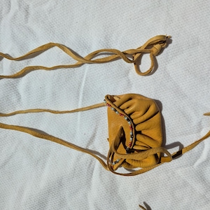 Puede incluir: Una peque&ntilde;a bolsa de cuero color canela con cierre de cord&oacute;n y adornos de cuentas. La bolsa es rectangular con bordes redondeados y est&aacute; conectada a una correa de cuero larga y delgada. La correa est&aacute; anudada en el extremo.