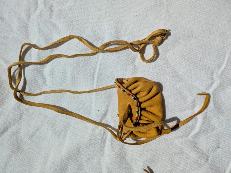 Puede incluir: Una peque&ntilde;a bolsa de cuero color canela con cierre de cord&oacute;n y adornos de cuentas. La bolsa es rectangular con bordes redondeados y est&aacute; conectada a una correa de cuero larga y delgada. La correa est&aacute; anudada en el extremo.