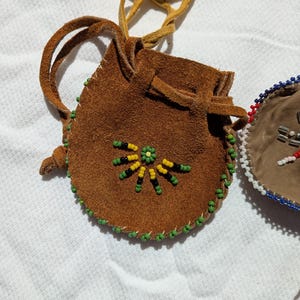 Puede incluir: Una peque&ntilde;a bolsa de gamuza marr&oacute;n con cierre de cord&oacute;n y un cord&oacute;n largo de cuero beige. La bolsa est&aacute; decorada con un dise&ntilde;o floral de cuentas amarillas, verdes y negras. Los bordes est&aacute;n adornados con cuentas verdes. Una segunda bolsa es visible en el fondo.