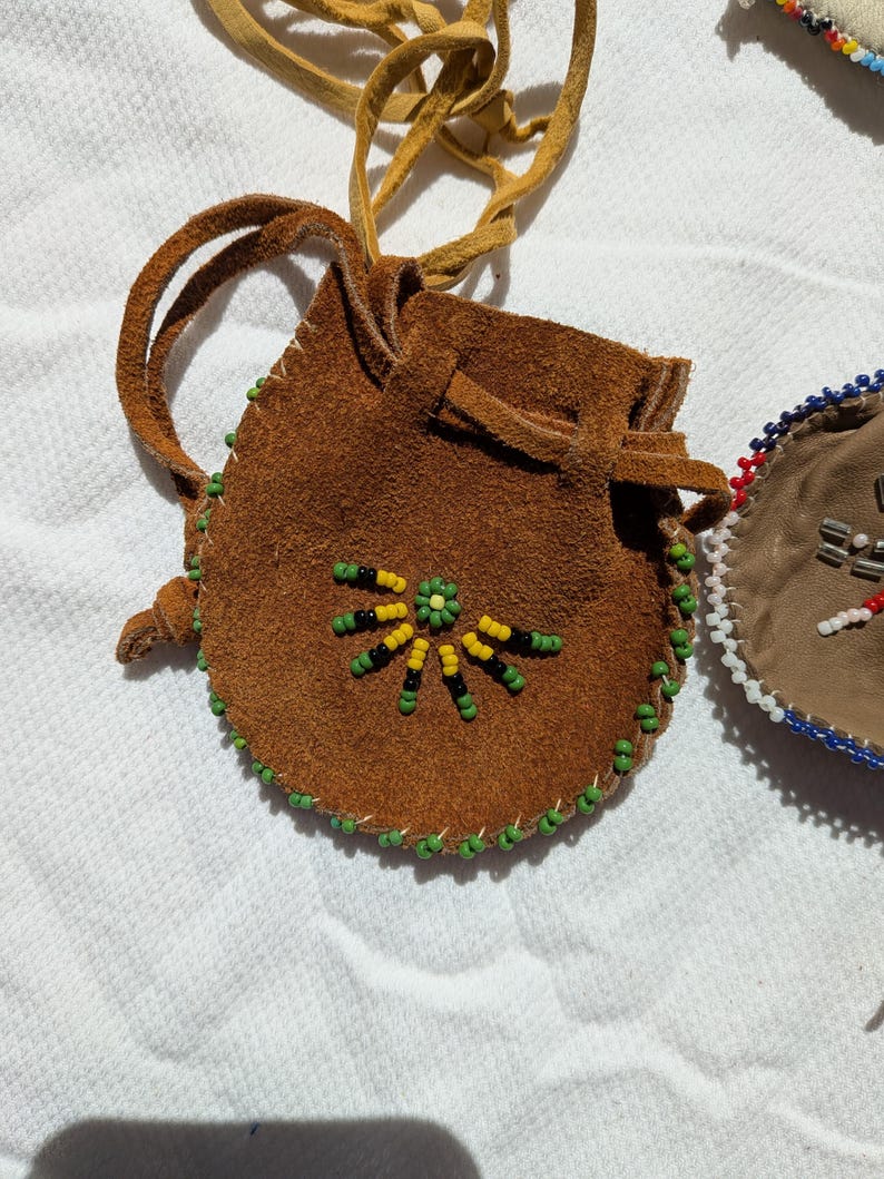 Puede incluir: Una peque&ntilde;a bolsa de gamuza marr&oacute;n con cierre de cord&oacute;n y un cord&oacute;n largo de cuero beige. La bolsa est&aacute; decorada con un dise&ntilde;o floral de cuentas amarillas, verdes y negras. Los bordes est&aacute;n adornados con cuentas verdes. Una segunda bolsa es visible en el fondo.