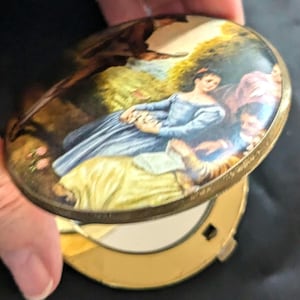 May include: A vintage gold-toned compact mirror with a hinged lid. The lid features a painted scene of three women in a garden setting. The women are wearing blue, yellow, and pink dresses. The background is a lush green forest.