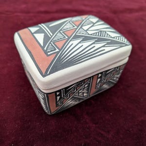 Pode incluir: Uma caixa retangular de cerâmica com tampa. A caixa é pintada com padrões geométricos em cores preto, branco e terracota. A tampa e as laterais apresentam desenhos intrincados, criando um recipiente de armazenamento decorativo.