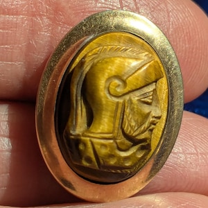 May include: Oval-shaped gold-tone brooch featuring a carved tiger's eye cameo of a man in a helmet. The cameo is set within a gold frame. The man's profile is detailed with a beard and helmet.
