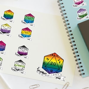 Pode incluir: Uma coleção de adesivos coloridos de dados D20 com o texto "HOW I ROLL" em uma faixa. Os dados apresentam vários esquemas de cores da bandeira do orgulho, incluindo o arco-íris, e são exibidos em uma superfície branca com uma caneta, clipes de papel e cadernos.
