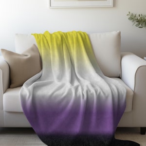 May include: A soft, yellow, white, purple, and black ombre throw blanket. The blanket is folded and draped over a white couch.