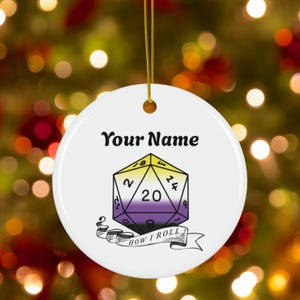 May include: White round ornament with a gold string for hanging. The ornament features a D20 die design in yellow, purple, and black, with the text "Your Name" above and "HOW I ROLL" below.