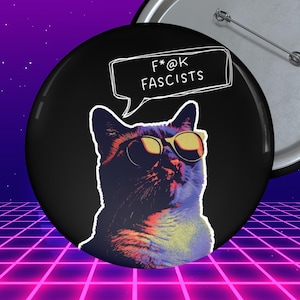 May include: Black button with a cat wearing sunglasses and a speech bubble that says "F*@k Fascists". The cat is a mix of purple, orange, and yellow. A second button is visible in the background.