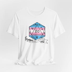 Peut inclure: T-shirt blanc avec un graphique d'un dé à vingt faces avec un schéma de couleurs du drapeau de la fierté transgenre. Le dé est entouré d'une bannière qui dit "How I Roll".
