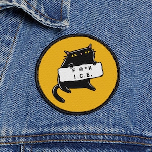 May include: A round patch featuring a black cat holding a sign with the text "F @*K I.C.E." The cat is set against a yellow background with a black border. The patch is on a blue denim jacket.