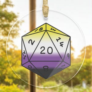 May include: A round, clear glass ornament with a 20-sided die printed on it. The die is colored in the colors of the non-binary pride flag: yellow, white, purple, and black. The die has the numbers 2, 4, 6, 8, 10, 12, 14, 16, 18, and 20 printed on it.