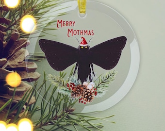 May include: A clear, round Christmas ornament with a black moth illustration wearing a Santa hat. The ornament has the text "MERRY MOTHMAS" in red. The moth is surrounded by pine needles, pine cones, and red berries.