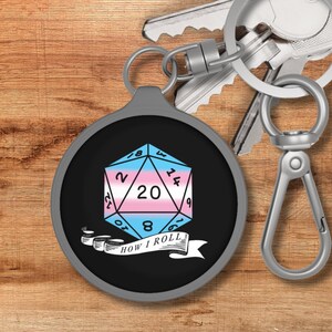 May include: A silver keychain with a black circle featuring a 20-sided die with a transgender pride flag design. The die has the numbers 2, 12, 20, 14, 6, 16, 8, 10, 18, 4, 9, 1, 3, 5, 7, 19, 11, 13, 15, and 17. Below the die is a banner that reads "How I Roll".