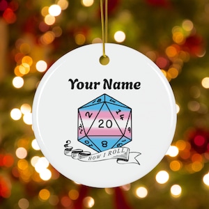 May include: White ceramic ornament with a gold string, featuring the text "Your Name" above a twenty-sided die with a transgender flag design. The phrase "HOW I ROLL" is written on a banner below the die.