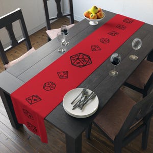 May include: Red table runner with black dice printed on it. The dice are different sizes and show different numbers. The runner is on a dark wood table with a plate and silverware.