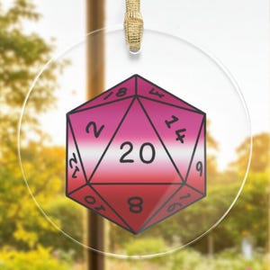 Könnte beinhalten: Ein transparenter Glas-Anhänger mit einem rosa und weißen 20-seitigen Würfel mit schwarzen Zahlen. Der Würfel ist eine Darstellung der lesbischen Pride-Flagge.