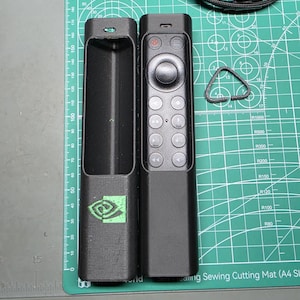May include: A black remote control case with a green Nvidia logo, a matching black remote control, and a small triangular black plastic component. The items are positioned on a green cutting mat with a visible ruler measuring up to 190 millimeters.