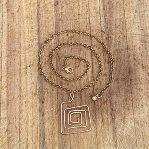 May include: A gold-colored necklace with a square pendant featuring an inner spiral design. The chain is a delicate link style, and the pendant is suspended from the chain. The necklace is displayed on a wooden surface.