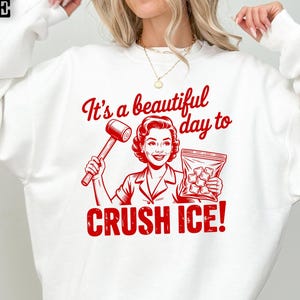 May include: White sweatshirt with a red graphic that reads "It's a beautiful day to CRUSH ICE!" The graphic features a vintage illustration of a woman holding a mallet and a bag of ice cubes.
