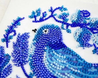 May include: A blue bird beaded with blue, light blue, and white beads, surrounded by blue beaded flowers on a white background.